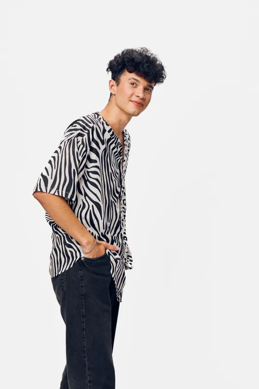 Young man wearing a zebra print shirt and black jeans stands casually with hands in pockets against white background, showcasing stylish streetwear and relaxed confidence.