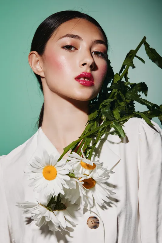 Elegant young woman with glossy red lips and radiant skin poses with vibrant white daisies in a creative fashion concept. Soft lighting highlights her swollen eyes and smooth complexion.