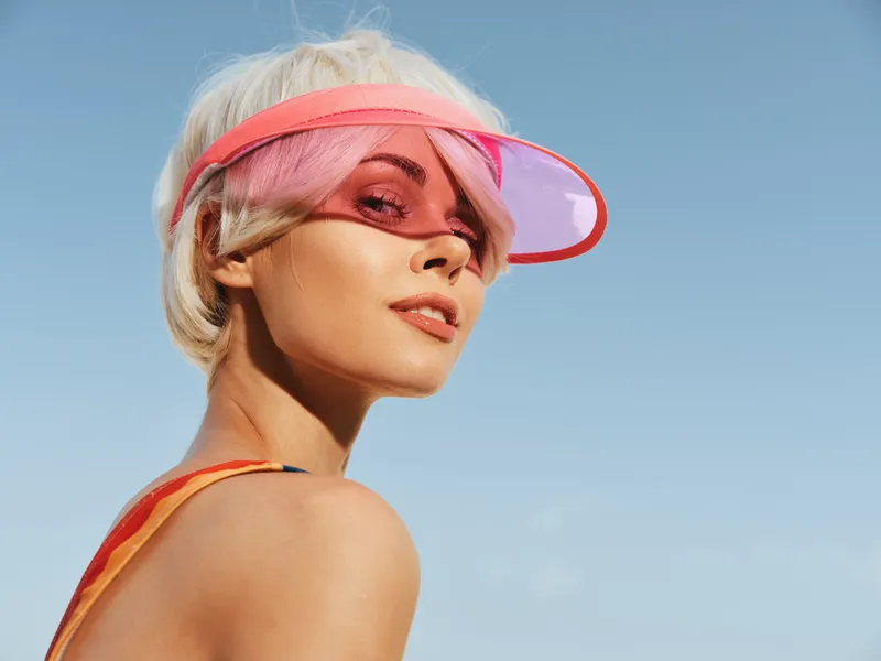 Portrait of a stylish woman wearing a pink sun visor against a clear blue sky, emphasizing contemporary beach fashion, sunny mood, and bold color contrast.