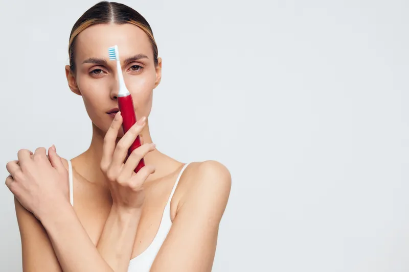 Young woman with clear skin holding red electric toothbrush near face, minimal makeup, neutral background, skincare concept, personal hygiene, beauty routine.