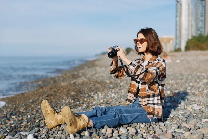 Woman photographer on a pebble beach wearing a plaid shirt, jeans and boots captures seaside scenery with a compact camera, sunglasses on, relaxed mood and ocean horizon in the background.
