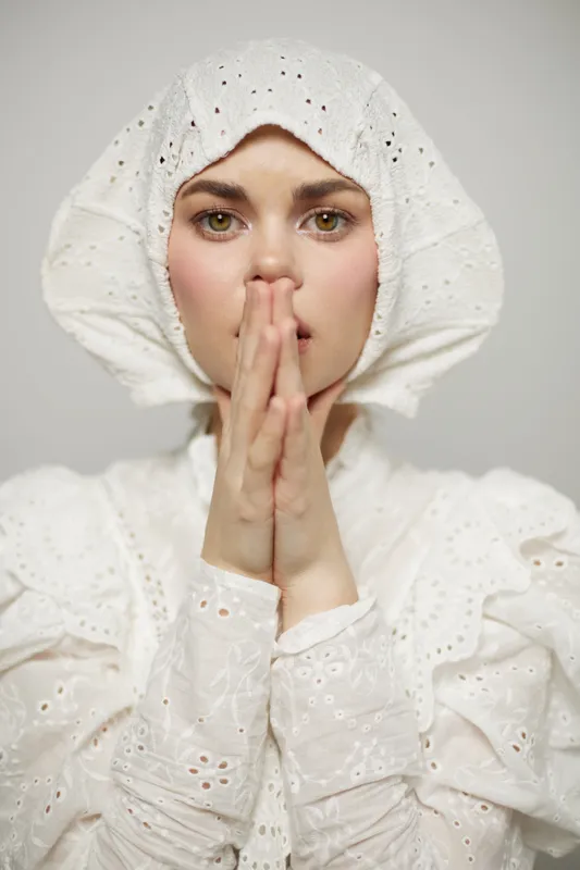 A woman with an emotional expression wears a white vintage bonnet and blouse, creating a serene atmosphere against a soft, neutral background