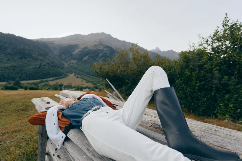 Woman relax on wooden bench in countryside meadow with mountains in background, wearing boots, white pants and denim jacket, outdoor leisure, rural escape and calm nature.