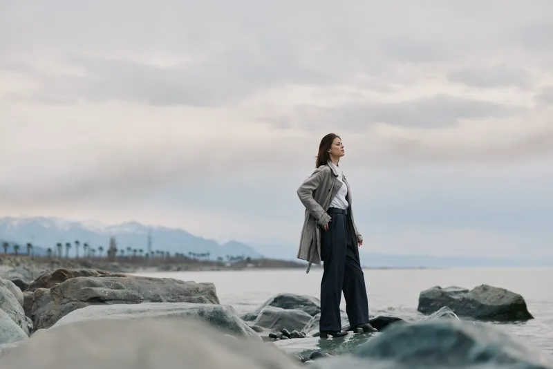 Young woman stands on rocky shore looking at horizon, dressed in casual clothes and coat. Calm sea and cloudy sky create serene mood for outdoor lifestyle and contemplation.