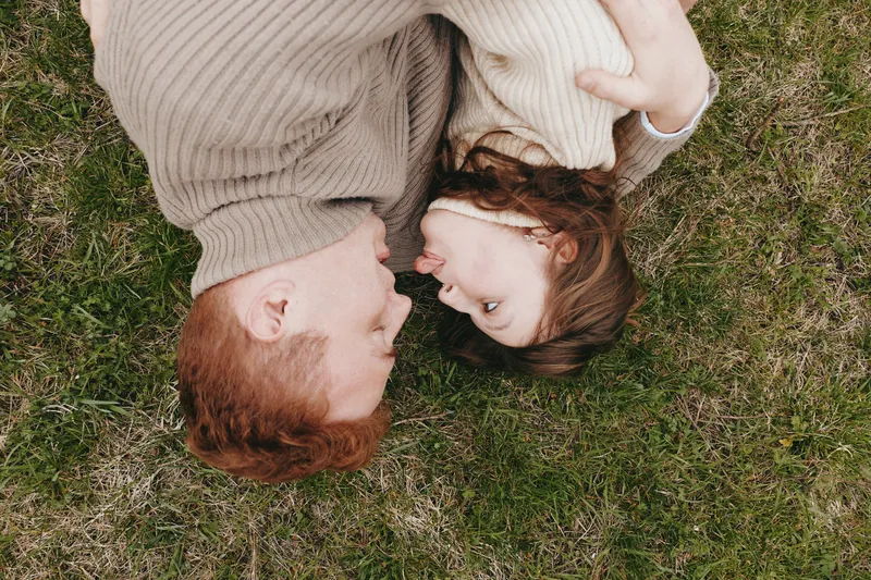 Young couple with red and brown hair lies closely together on green grass sharing an intimate moment in casual sweaters, conveying love and connection in a natural lifestyle setting.