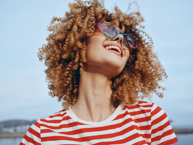 Vibrant portrait of a joyful woman with voluminous curly hair, wearing colorful sunglasses and a striped tee, basking in bright outdoor light with carefree energy, against a clear sky, capturing