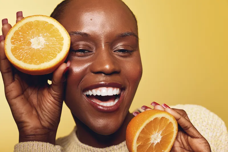 Happy African woman with dark skin smiling and holding two orange halves near face on yellow background. Concept of healthy lifestyle, vitamin c, and natural beauty.