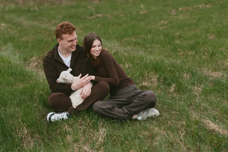 A couple sits on grass with a goat kid nearby, outdoors in a field, sharing a warm moment. Lively, wholesome lifestyle scene highlighting love, bonding, nature, and relaxed countryside atmosphere.