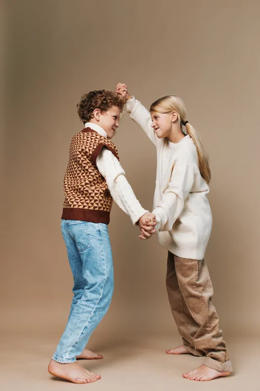 Authentic moment of children at play, two kids hold hands and dance in a warm studio mood, casual school inspired fashion with soft textures and cozy layers.
