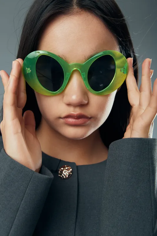 Young woman wearing large green sunglasses with dark lenses and long dark hair in gray clothing, portrait with serious expression and hands adjusting glasses indoors on gray background.