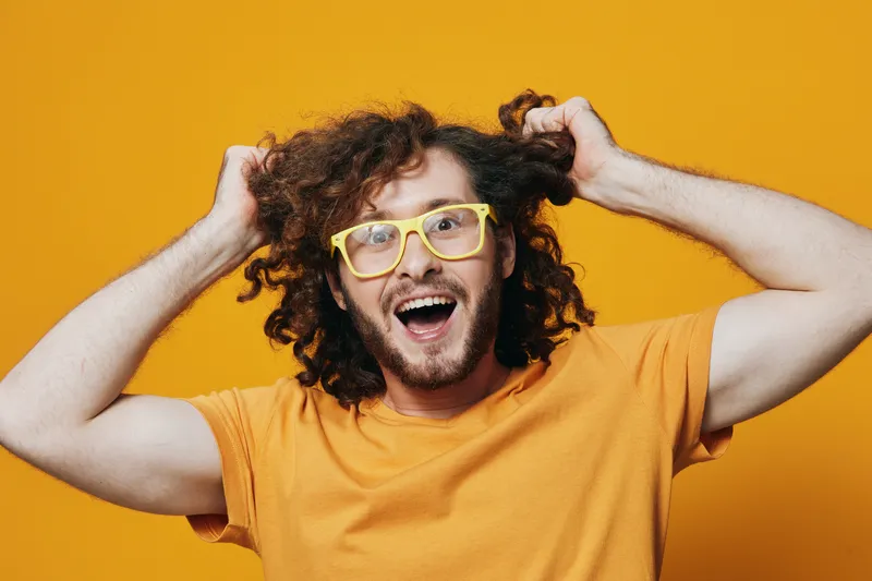 Cheerful young man with curly hair wearing bright yellow glasses and an orange t shirt, expressing excitement against a vivid yellow background