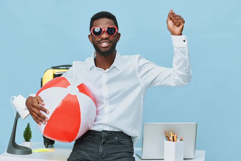 Happy cheerful young man with sunglasses holding beach ball and travel bag, dressed casually in white shirt, celebrating vacation, in bright studio with blue background. People lifestyle concept, summ
