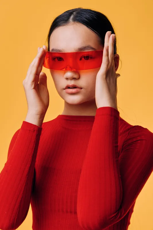 Young woman in red sweater wearing futuristic red sunglasses with hands adjusting eyewear on orange background. Portrait of stylish girl with sleek hair and modern fashion accessories.