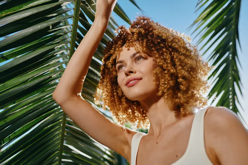 smiling woman with curly hair enjoying sunlight under green palm leaves on a bright summer day, natural beauty, tropical vibes, warm atmosphere