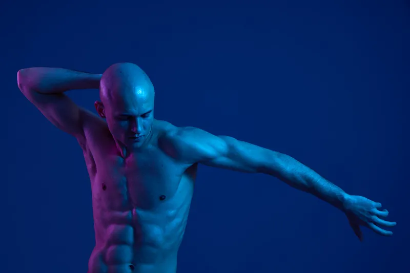 Male model posing dynamically against a blue background, showcasing a strong physique and expressive body language, emphasizing creativity and movement in art.