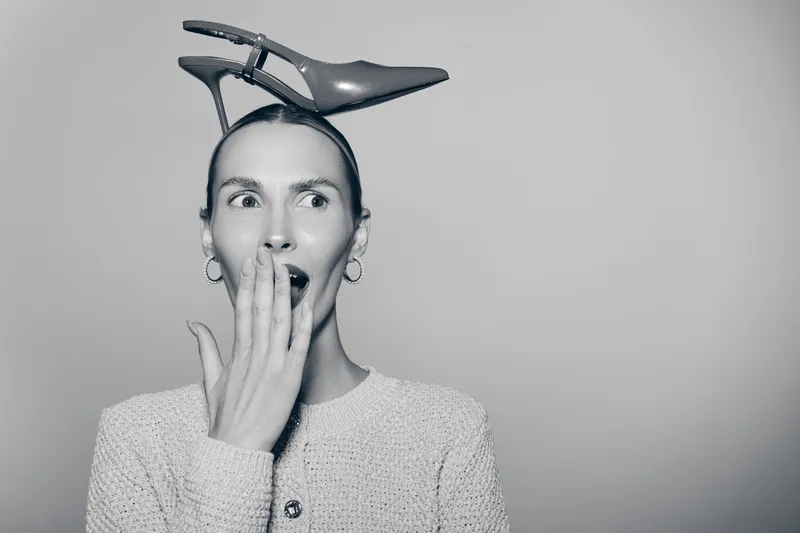 Surprised white woman with light skin and high cheekbones holds hand near mouth with shoe balanced on her head in studio.