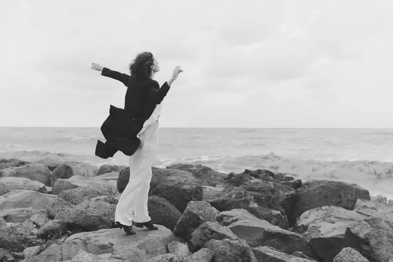 Candid couple dances on rugged rocks beside the sea, capturing motion and joy against a vast ocean horizon, wind lifting garments in a dramatic monochrome scene
