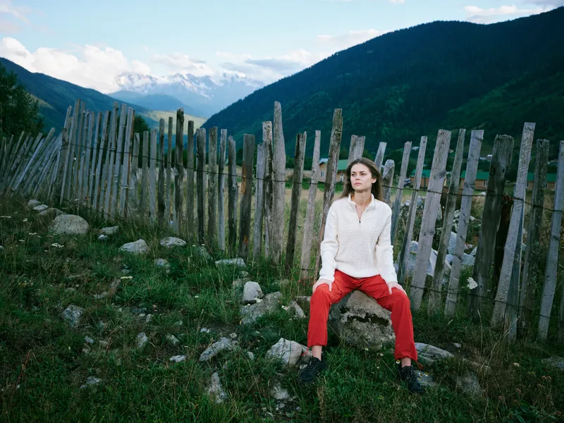 Woman sits beside a weathered wooden fence in a mountainous landscape, wearing red pants and a light sweater, with green grass, rocks, and distant snowcapped peaks.