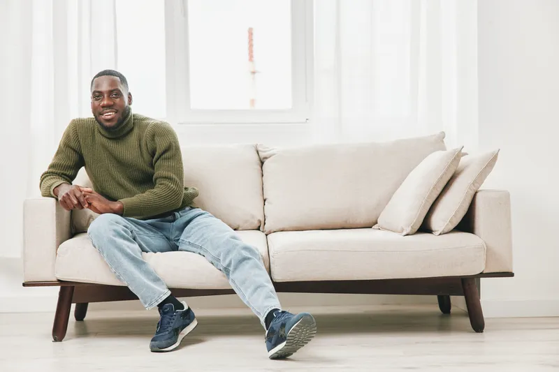 Smiling man sitting on a sofa in a stylish living room, wearing a green sweater and jeans, conveying warmth and comfort, ideal for lifestyle themes