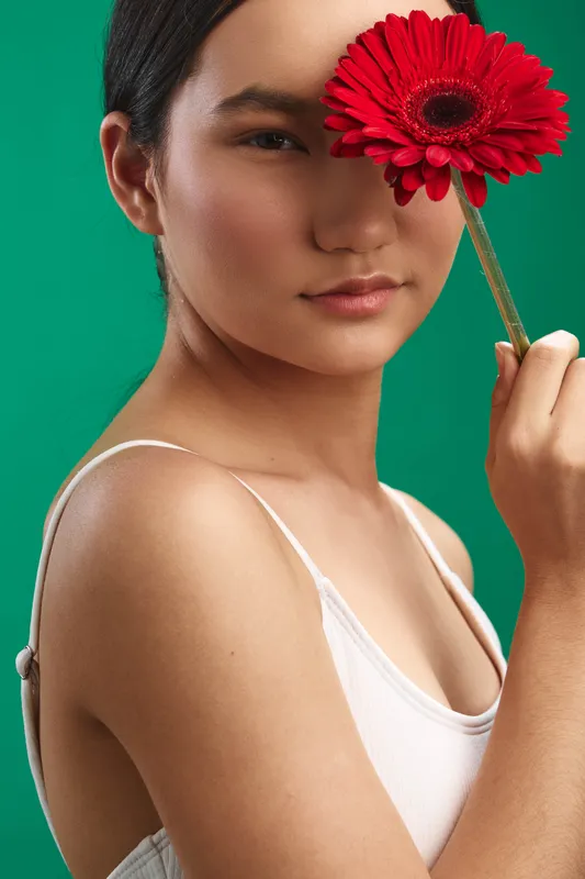 Asian woman gazes softly at the camera while holding a red flower near her face, conveying elegance, calm confidence, and a vibrant studio mood against a green backdrop.