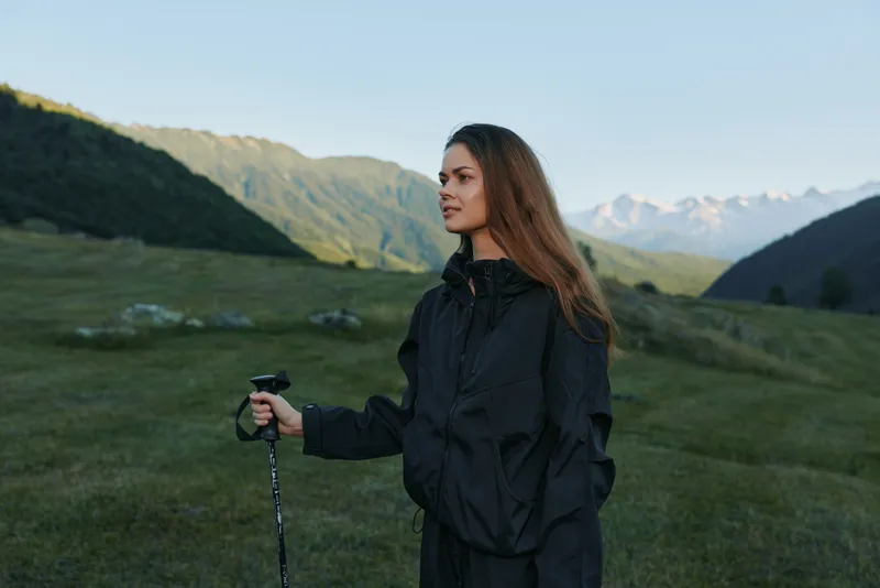 A lone female hiker stands in a green alpine meadow with a trekking pole. She wears a dark jacket and looks toward the distant hills, calm and composed in wide open nature.