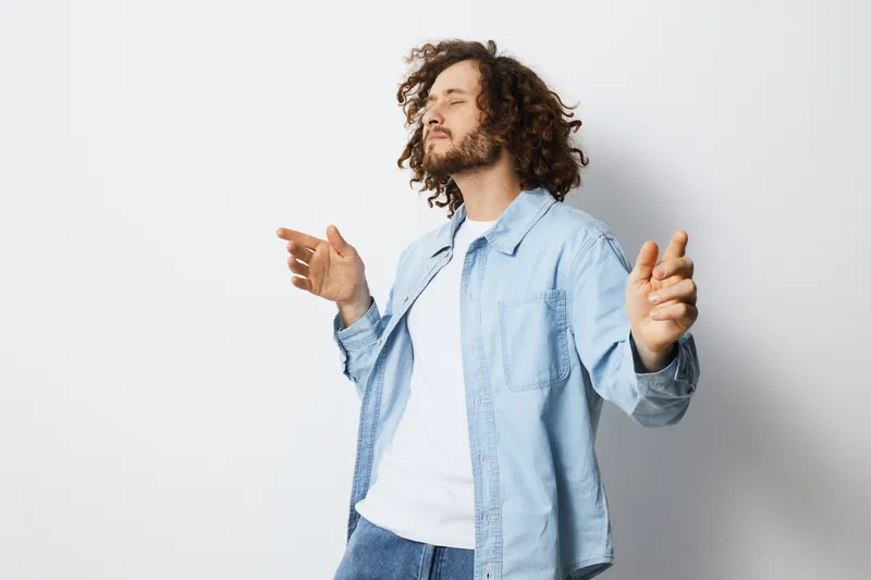 Young man with curly hair enjoying music indoors, wearing a denim jacket, expressing joy against a minimalistic white background, perfect for lifestyle concepts