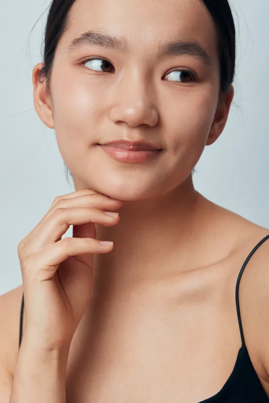 Young woman with bare shoulders and natural makeup looks sideways with gentle smile and hand near face against plain background, portrait of serene beauty and confidence.