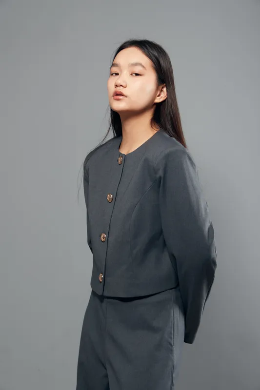 Young woman wearing gray formal suit with buttons posing with hands behind back on gray background. Asian female model with long dark hair in business style outfit for fashion portrait.