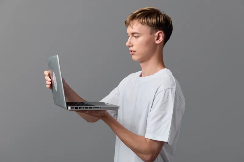 Laptop evaluation by a slim man in a plain white tee, holding the device with one hand and studying the screen, minimal studio setup, tech workflow concept