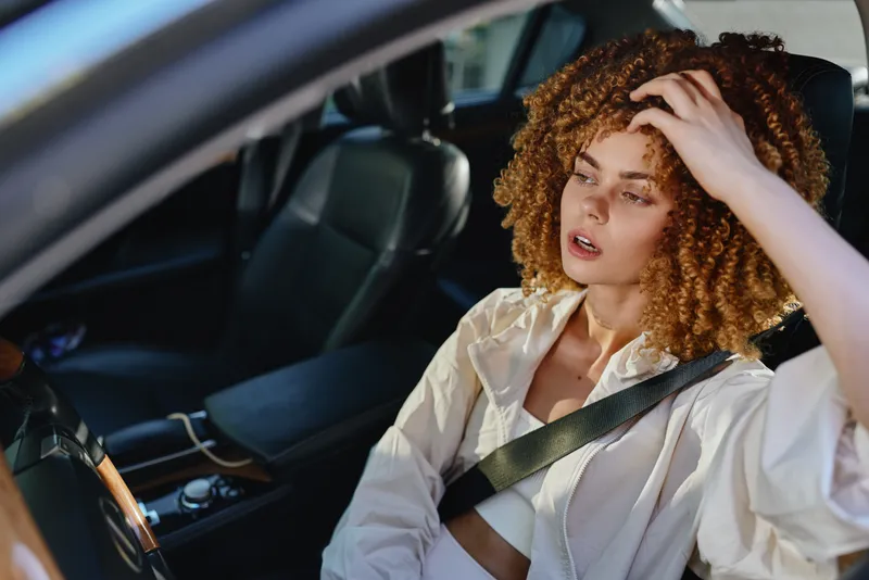 thoughtful woman with curly hair sitting in a car wearing a seatbelt, natural light highlighting her face, casual jacket, introspective mood captured