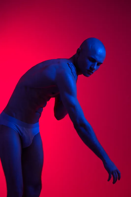 Slim man posing against a vibrant red and blue background, showcasing his physique with a dynamic gesture. The lighting enhances the artistic composition of the image.