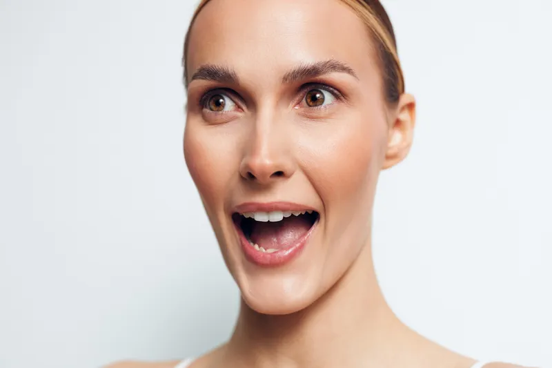 Surprised young woman with open mouth and wide eyes looks excitedly to the side against white background, showing natural skin and expressive facial expression.