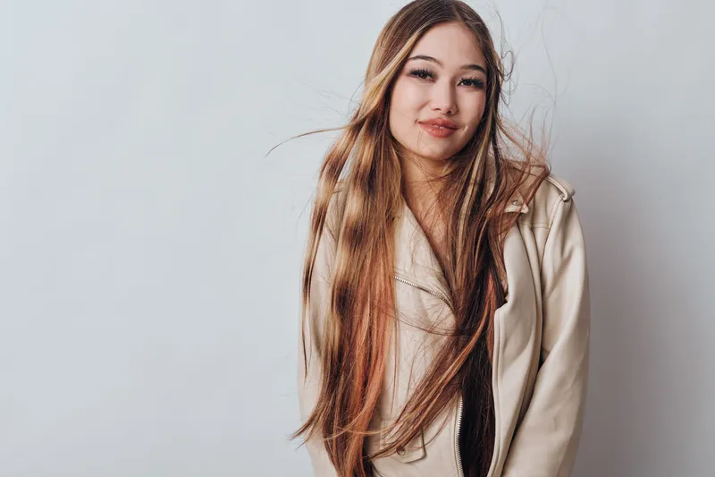 Young woman with long hair and natural makeup posing indoors against plain background, wearing beige jacket for casual fashion and beauty portrait session.