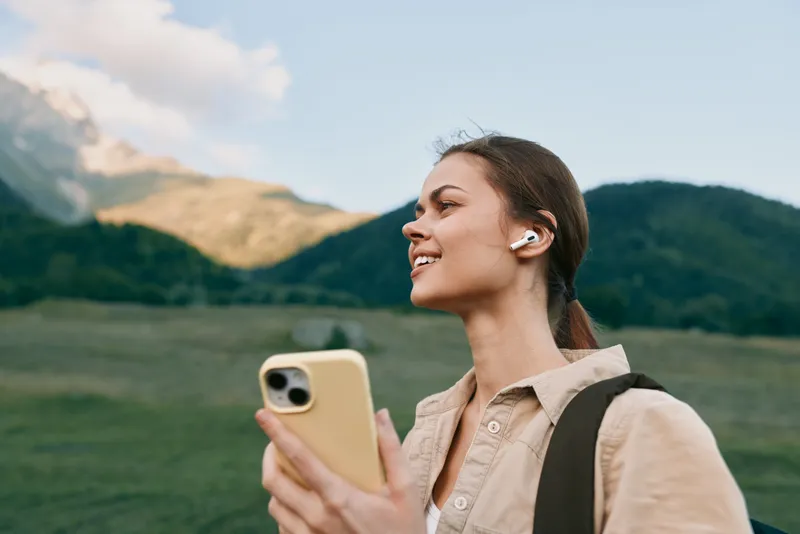 Woman outdoors using smartphone, smiling, wearing beige shirt and wireless earbuds, with backpack, open field and mountains in the background