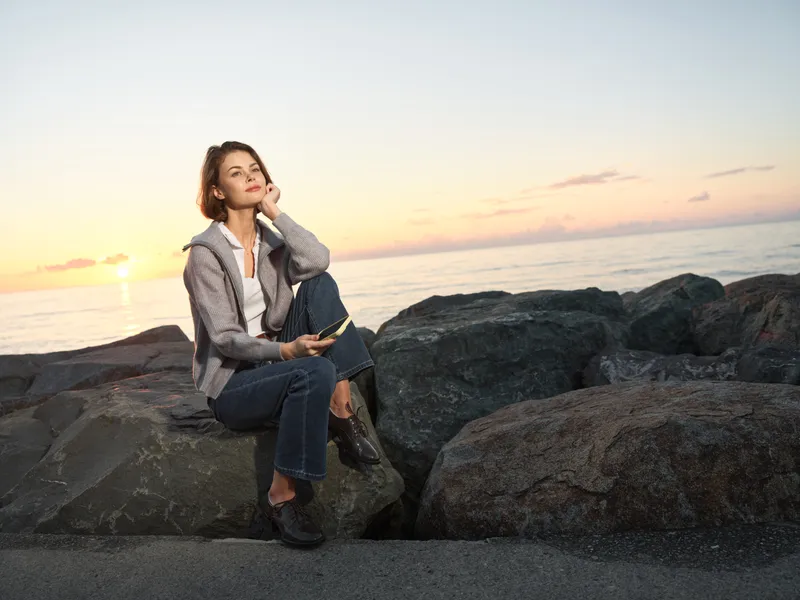 A thoughtful woman sits on a rocky shore at sunset, gazing into the distance with a notebook. She wears casual clothing and a gentle smile, creating a serene, reflective mood.