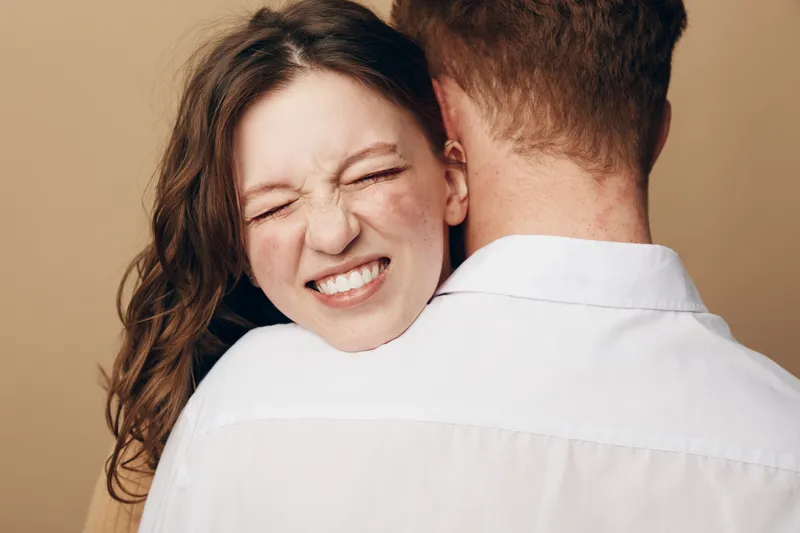 Young woman with closed eyes and smiling face hugs man wearing white shirt from behind. Intimate moment, affection, love, and connection between couple in warm studio setting with beige background.