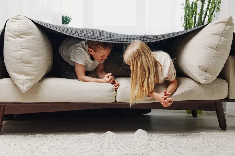 Two playful children hide under a gray blanket on a beige modern sofa in a bright living room. Concept of childhood fun, friendship, and indoor games.