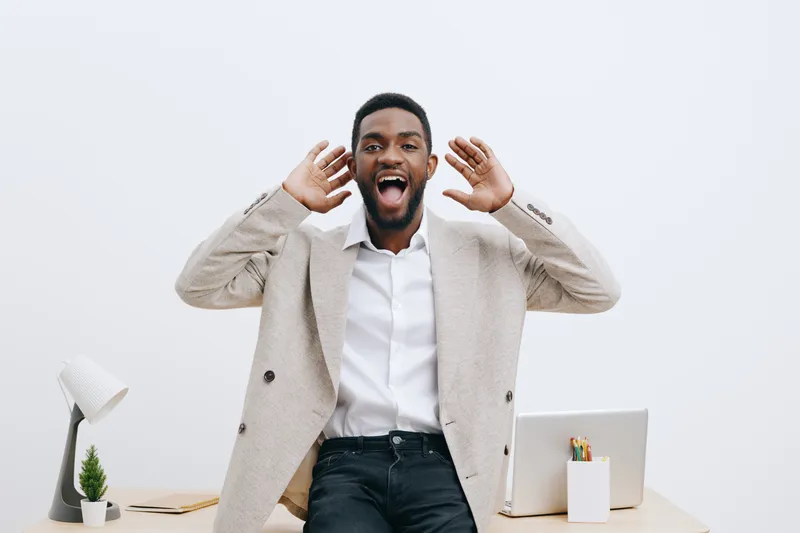 Happy smiling man with dark skin, casual beige blazer and white shirt, gesturing excitedly near office desk, isolated on plain white background. People lifestyle concept, cheerful, professional, posit