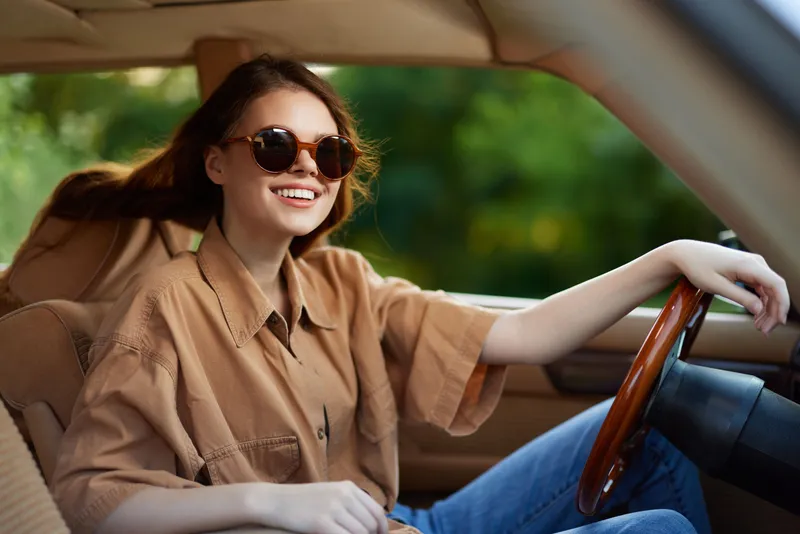 Young woman smiling in a stylish oversized shirt and sunglasses, enjoying a drive in a vintage car surrounded by lush greenery, conveying a carefree summer vibe.