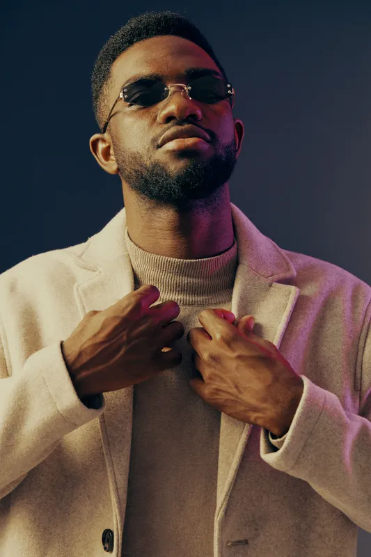 Stylish African man posing confidently in sunglasses and a beige coat against a colorful background, capturing contemporary fashion and self assurance