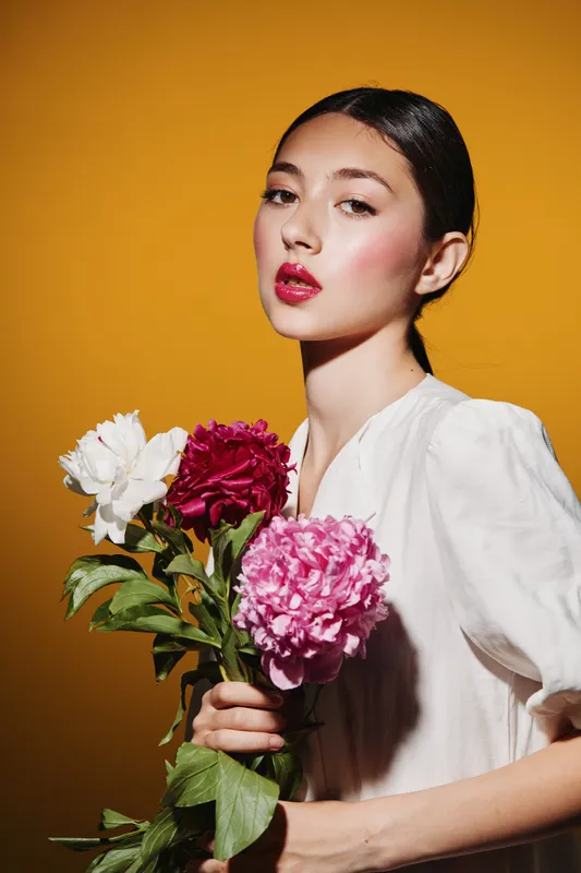Elegant young woman with glowing skin and red glossy lips holds colorful flowers. Bright fashion beauty portrait on warm yellow background representing creative style.