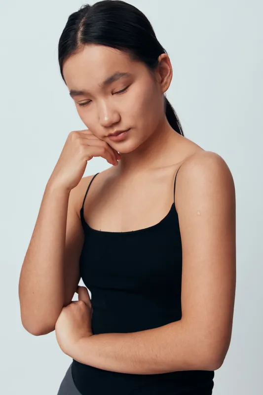 Young woman with long dark hair stands with eyes closed and hand on chin wearing black tank top. Minimalist background and natural skin tone, thoughtful pose with calm expression.