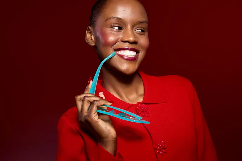 Smiling black woman with dark skin wearing a red sweater holds turquoise glasses near her mouth. Studio portrait on red background with bright lighting and positive mood concept.