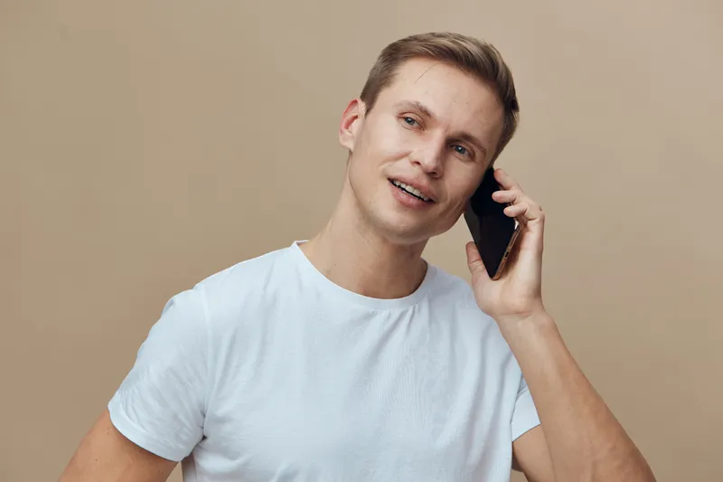Male young adult smiling man wearing casual white t-shirt talking on mobile phone isolated on neutral beige background studio portrait. People lifestyle concept