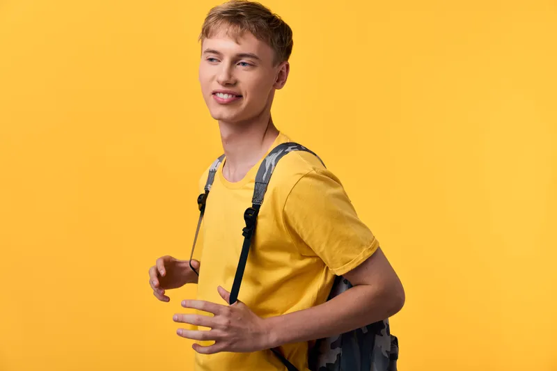 Young man with backpack wearing a yellow t shirt stands against a bright backdrop, casual student style conveying energy, confidence, and readiness for class, travel, or new experiences.