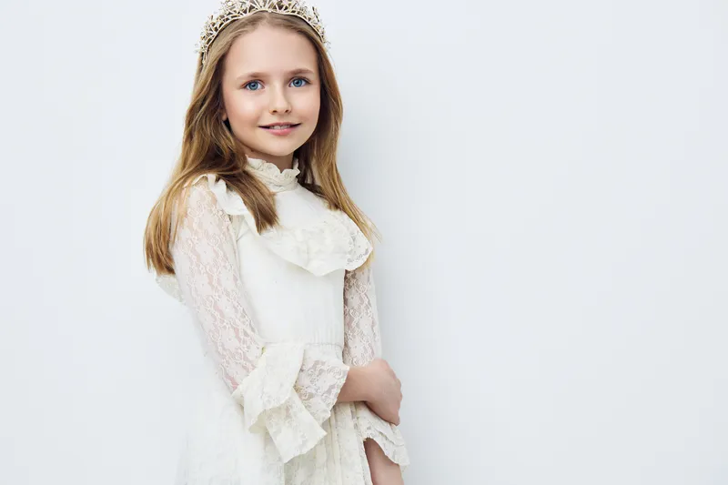 Cheerful young girl wearing a white lace dress and a crown, smiling gently against a light background, showcasing innocence and elegance