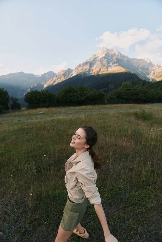 Young woman enjoying nature in a green field with mountains in the background. Lifestyle concept with fresh air, freedom, and outdoor adventure on a sunny day.