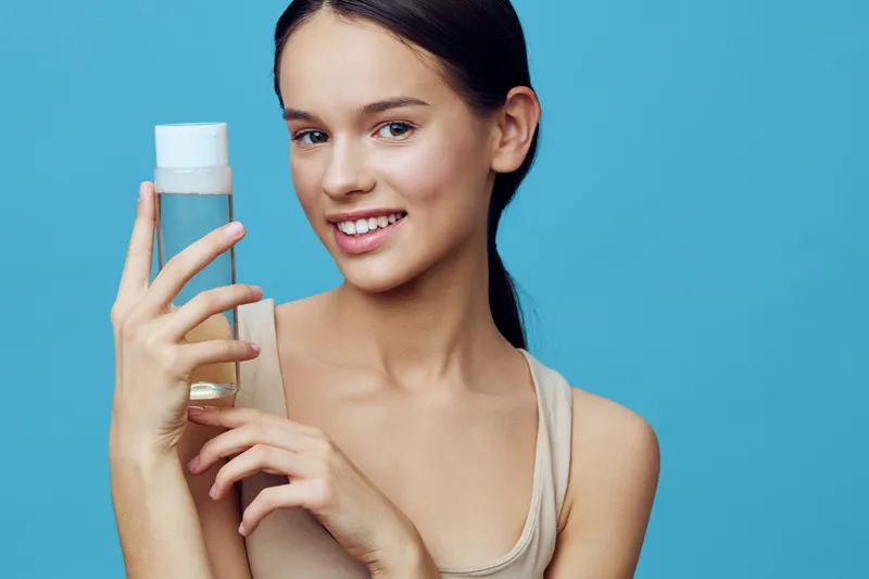 Young woman holding a skincare bottle, smiling against a blue background, showcasing a healthy skincare routine for glowing skin