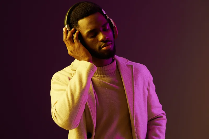 Young Black man in stylish attire, enjoying music with headphones against a vibrant purple background, exuding relaxation and joy through emotional expression