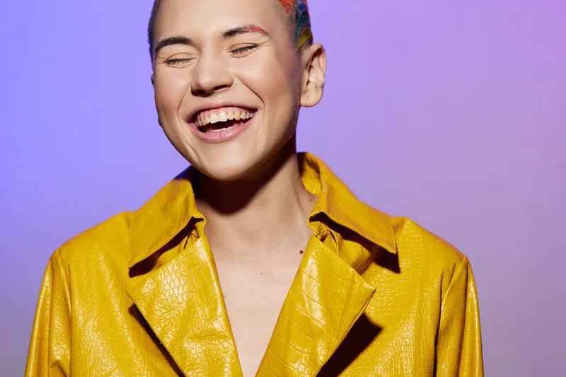 Smiling woman with colorful hair and yellow textured jacket laughing joyfully against purple background, expressing happiness and vibrant personality.